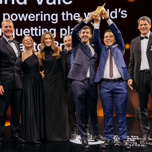 A group of people in formal attire stand on stage, two holding up a trophy, celebrating an award, with a presentation screen in the background.