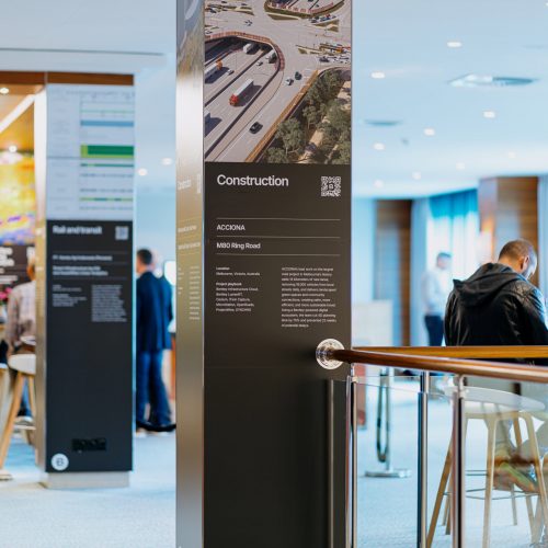 Indoor event space with informational panels on construction, people walking and sitting, and a railing in the foreground.