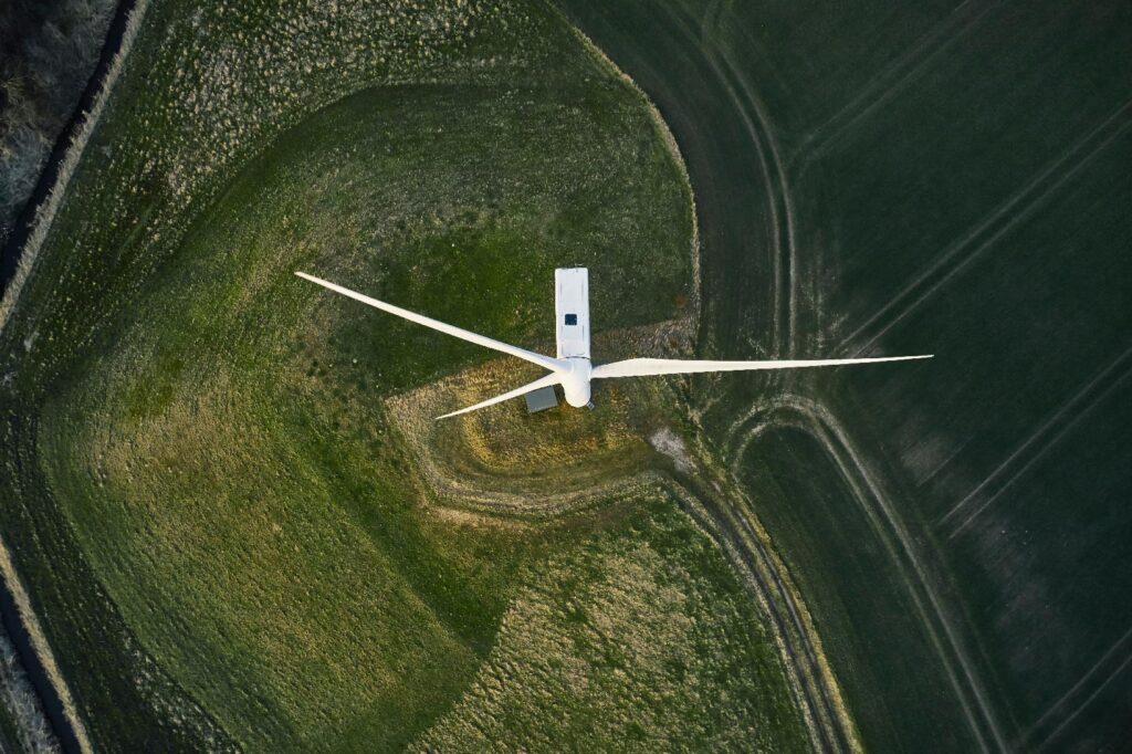 Aerial view of a white wind turbine surrounded by green fields with visible paths and patches of grass.
