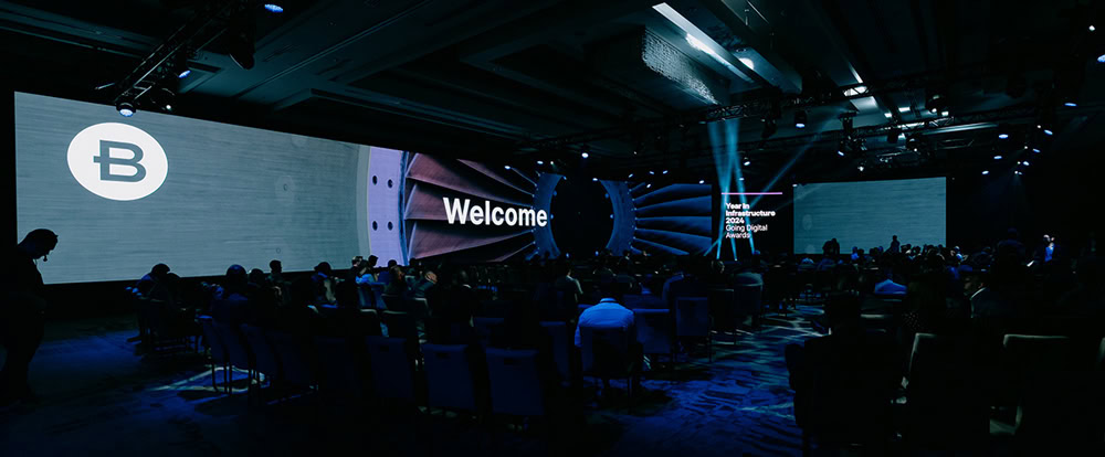 A large conference room with people seated, facing a stage with wide screens displaying "Welcome" and a circular logo on the left. Blue lighting fills the space.