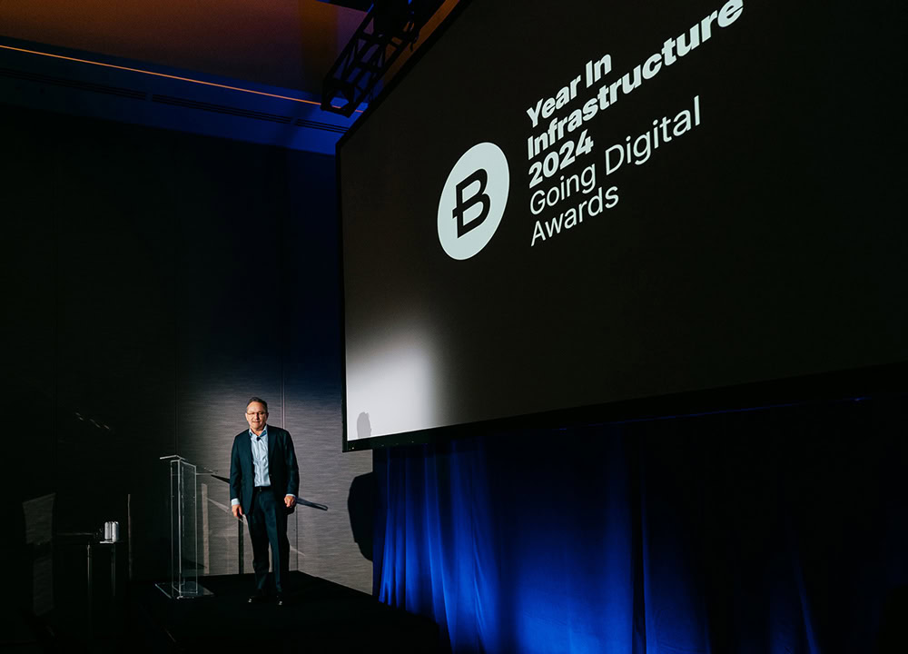 A man stands on stage next to a large screen displaying "Year in Infrastructure 2024 Going Digital Awards.