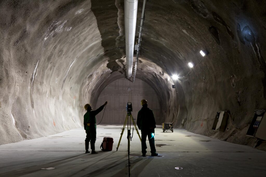 Two workers wearing hard hats stand in a large, empty underground tunnel, operating surveying equipment under ceiling lights.