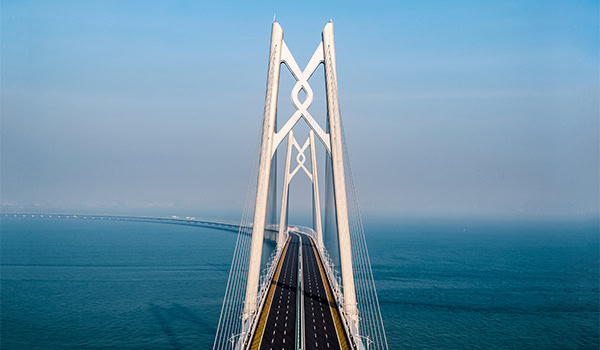 Aerial view of a cable-stayed bridge extending over a calm blue sea, with clear skies in the background.