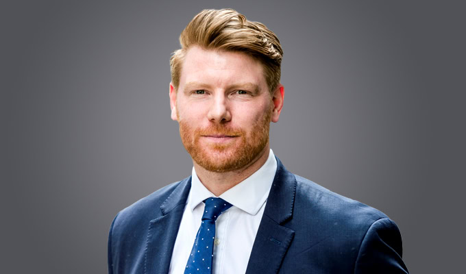 Guy Beaumont, a man with light skin, reddish hair, and a beard, is wearing a blue suit, white shirt, and blue tie while standing against a plain grey background.