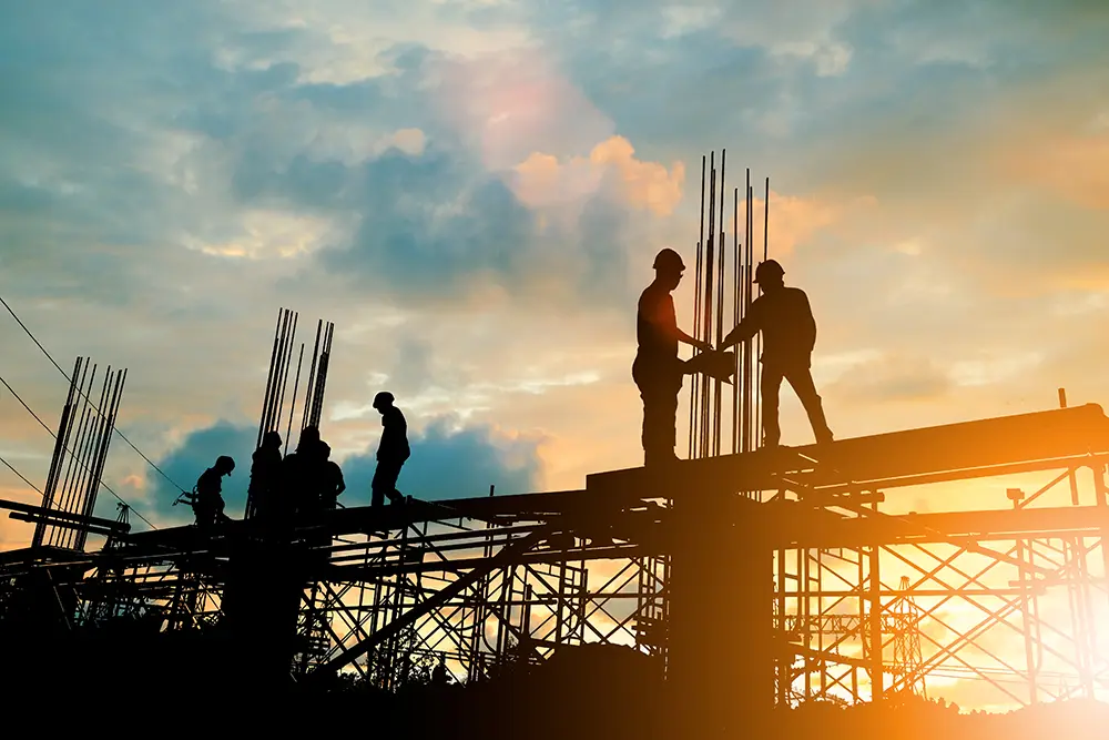 Silhouetted construction workers on scaffolding at sunset, handling metal rods and working together against a cloudy sky, embodying the strength and precision often guided by expert advisory services.
