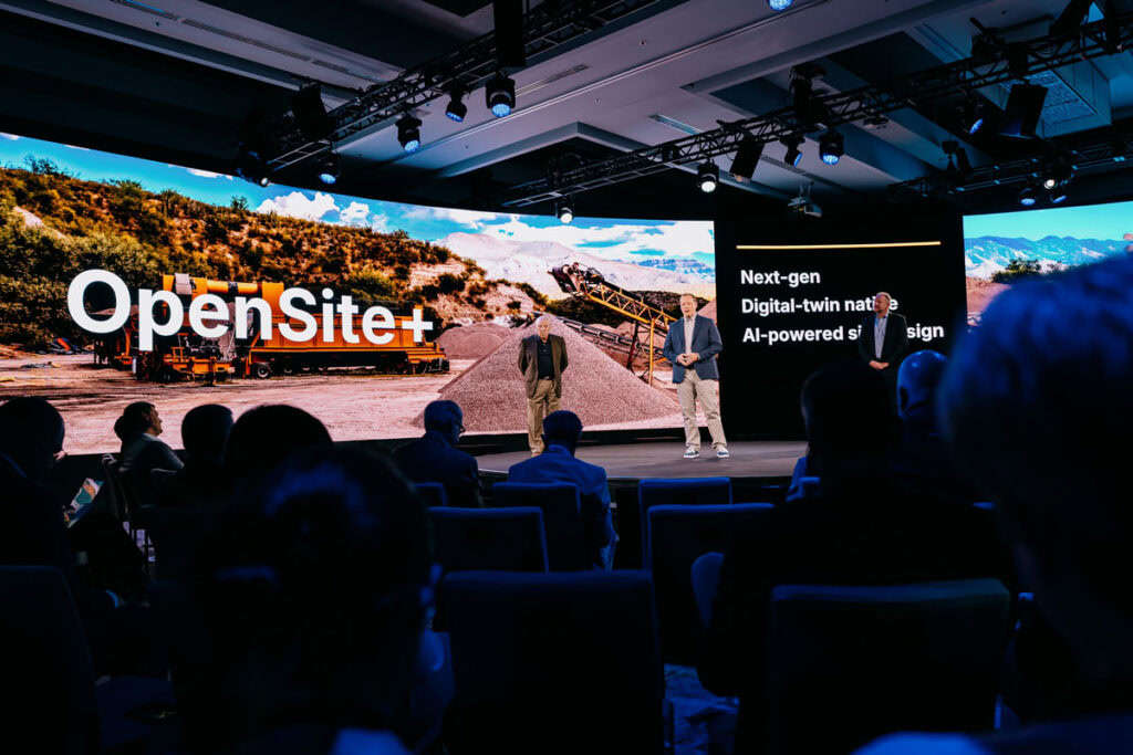 Dois apresentadores estão no palco em uma conferência de tecnologia, com o "OpenSite+" e a tecnologia de gêmeos digitais destacadas em telas grandes atrás deles.