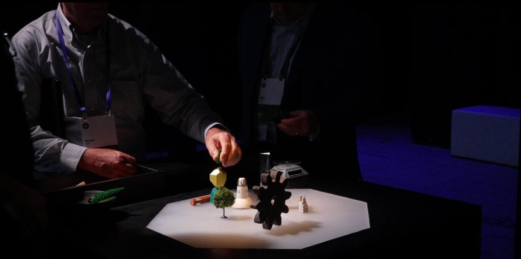 Two people interact with small models, including a tree, a lightbulb, and a gear, placed on an illuminated tabletop in a dark room.