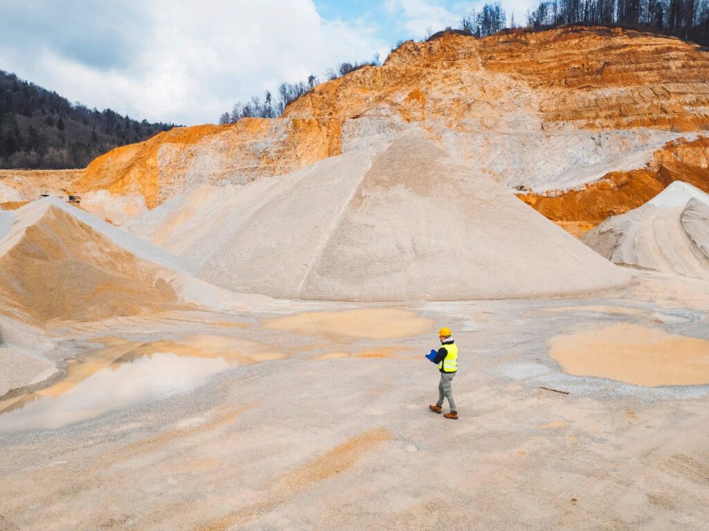 A person in a high-visibility vest and helmet walks across a large open-pit quarry with piles of sand and gravel, surrounded by rocky terrain and some puddles.