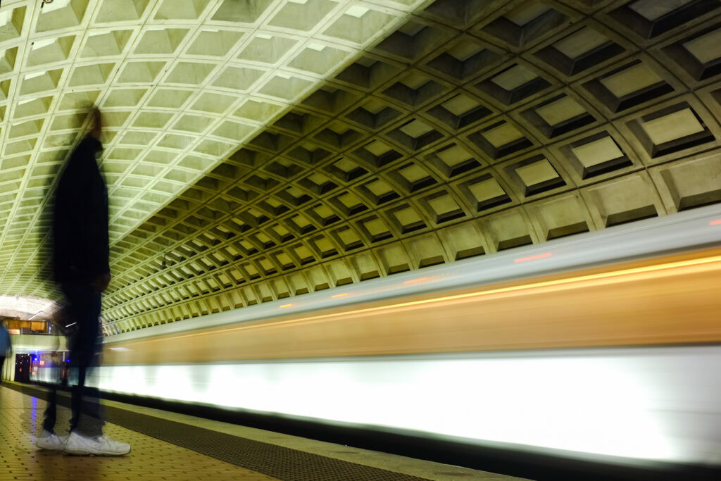 A person stands still on a subway platform as a train speeds by, captured in motion blur, under a geometric concrete ceiling.