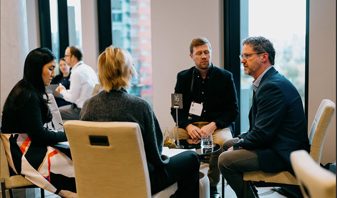 Four people sit around a small table in a modern office space, engaged in conversation, with city buildings visible through large windows in the background.