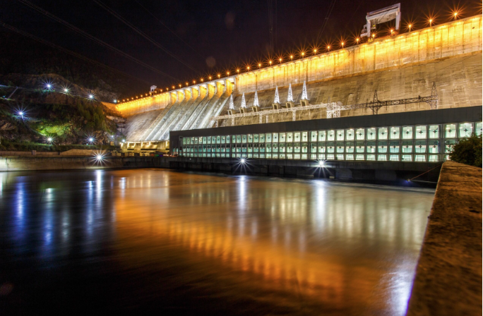 A large dam is illuminated at night with bright lights reflecting on the calm water below, as detailed in the CMR Dam Report. The structure features multiple sections and a row of lights near the top.