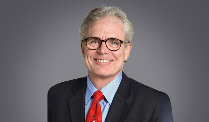 A man with gray hair and glasses, wearing a dark suit, light blue shirt, and red tie, smiles at the camera against a plain gray background.