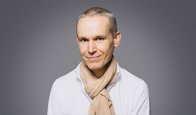 Julien Moutte, a man with short gray hair, wears a white shirt and a beige scarf, standing against a plain gray background.