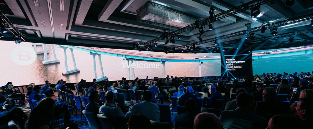 A large conference hall filled with seated attendees facing a wide stage with digital screens displaying "Welcome" and event information.