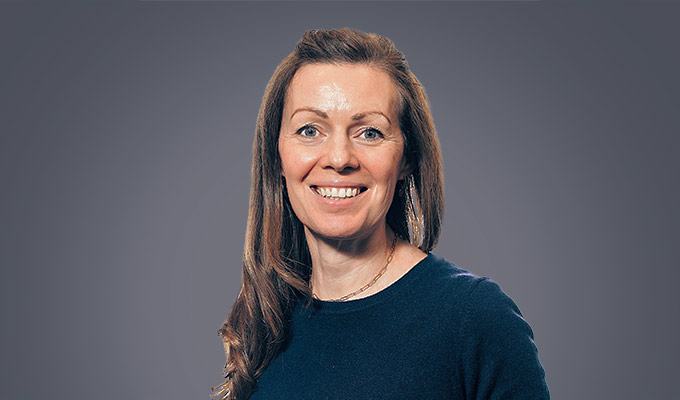 A woman with long brown hair wearing a navy blue top smiles at the camera against a plain gray background.