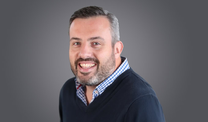 A man with short dark hair and a beard is smiling, wearing a dark sweater over a blue checkered shirt, posed in front of a plain gray background.