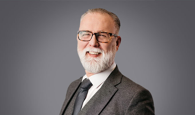 A middle-aged man with gray hair, a white beard, and glasses, wearing a gray suit and tie, smiles against a plain gray background.