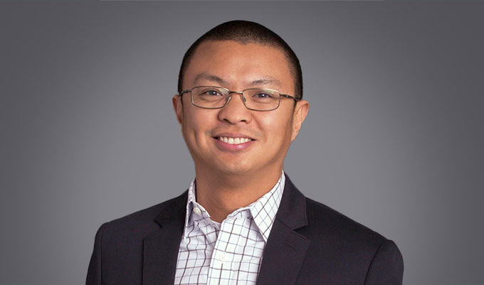 Mark Tablante, wearing glasses, a dark blazer, and a checkered shirt, is smiling in front of a plain gray background.