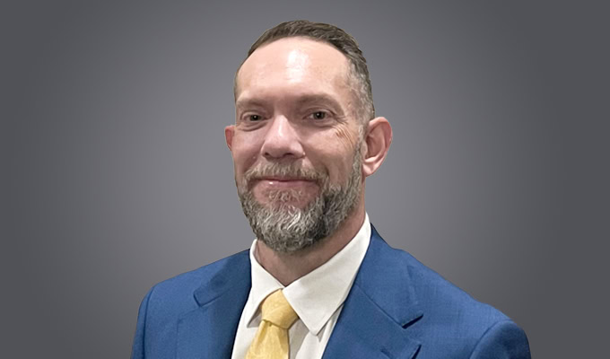 Michael Pearson, a man with short hair and a beard, wearing a blue suit, white shirt, and yellow tie, poses in front of a gray background.