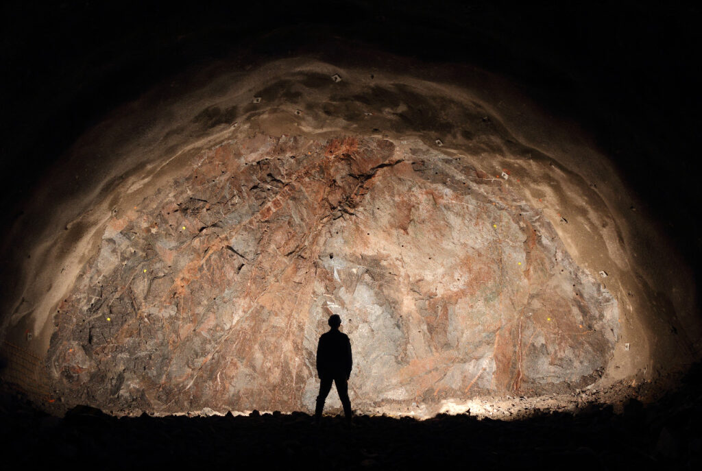 A person stands silhouetted in front of a large, illuminated rocky wall in a dark cave or underground tunnel.