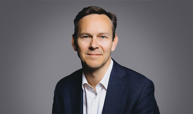 Nicholas Cumins, dressed in a dark suit and white shirt, sits and smiles at the camera in front of a plain grey background.