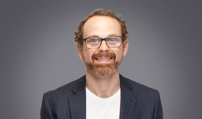 Patrick Cozzi, a man with glasses, a beard, and curly hair, is wearing a dark blazer over a white shirt and stands in front of a plain gray background.
