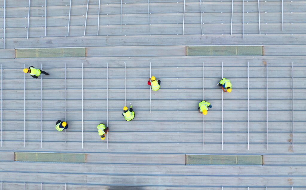 Aerial view of construction workers in yellow safety gear positioning metal bars on a large gray rooftop.