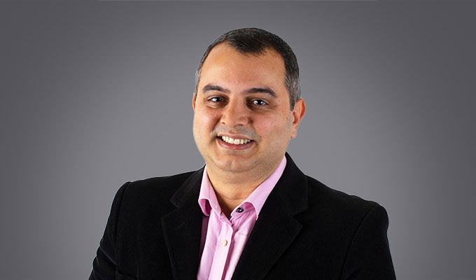 Rafael de Santiago Silva, dressed in a black blazer and pink shirt, smiles at the camera against a plain grey background.