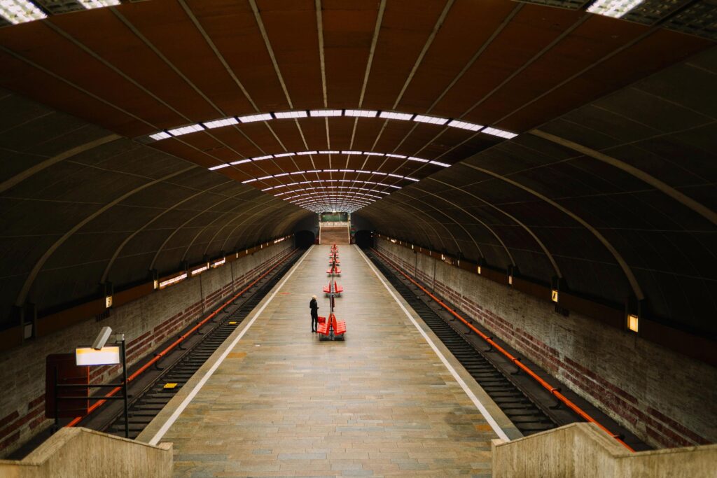 A person stands on an empty, covered subway platform lined with orange traffic cones, with train tracks on either side and no trains present.