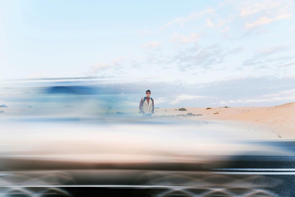 A person stands in a desert landscape as a blurred car speeds by on the road in the foreground under a partly cloudy sky.