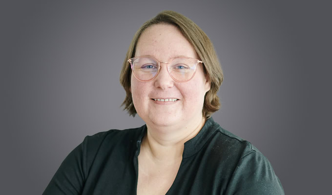 Savannah Altvater, with short brown hair and glasses, is smiling at the camera, wearing a dark top against a plain gray background.