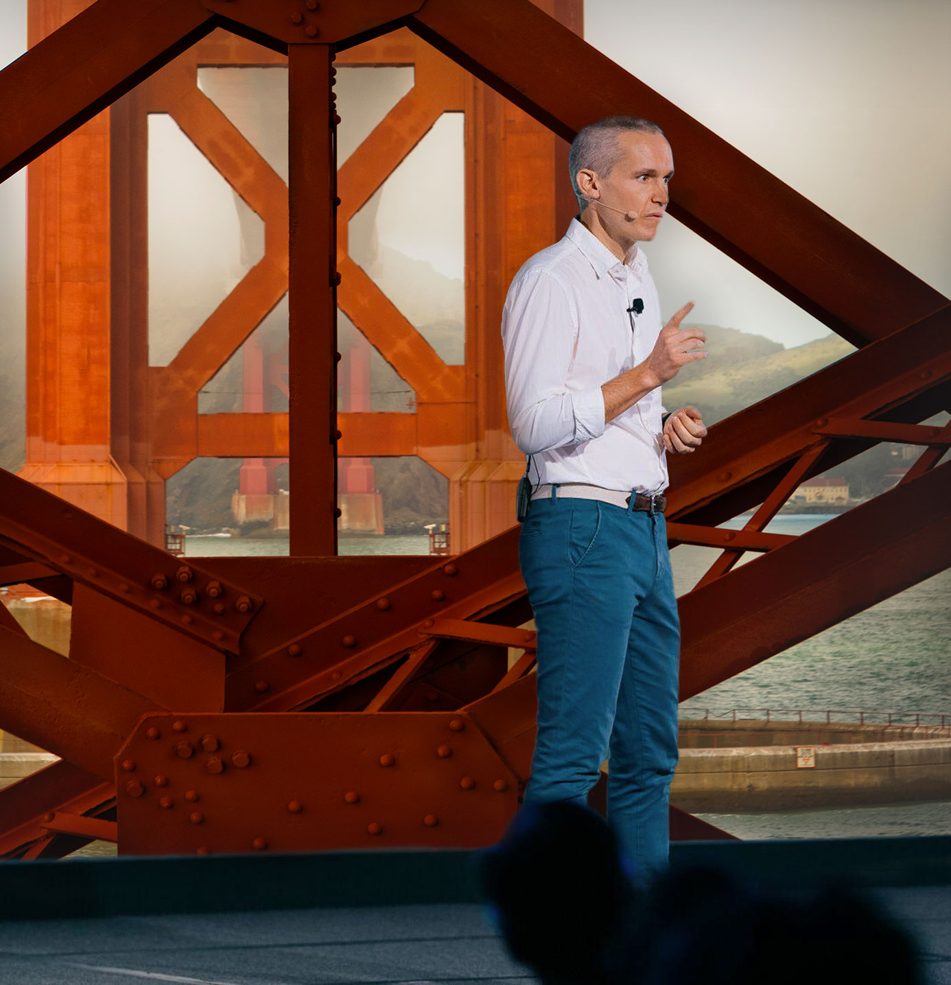 A man in a white shirt and blue pants stands and speaks into a microphone, outlining the agenda with a backdrop resembling the structure of a large bridge, possibly the Golden Gate Bridge.