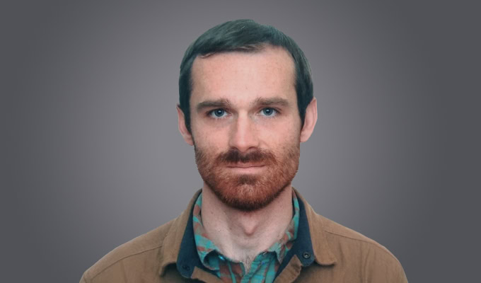 A man with short brown hair, a beard, and mustache is facing forward, wearing a brown jacket over a green and brown patterned shirt, set against a plain grey background.