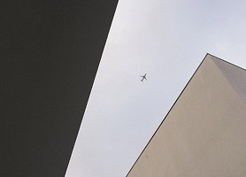 A commercial airplane is seen flying in the sky, framed by the angular edges of two modern buildings.