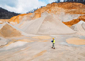 A worker in a yellow vest and helmet stands in a large open-pit quarry surrounded by piles of sand and gravel, with exposed earth and hills in the background.