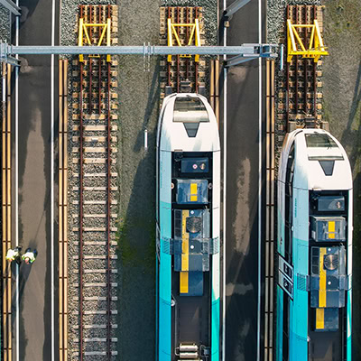 Aerial view of a railway yard in Q4, with two trains parked on tracks. Two people in safety gear are walking beside the trains.