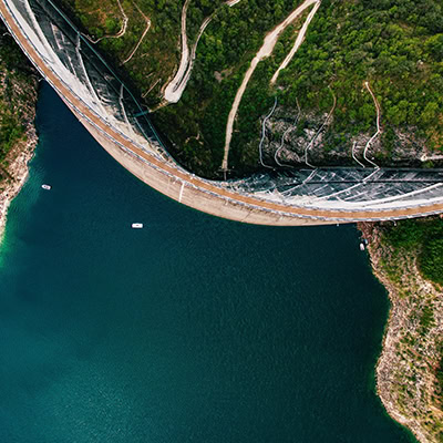 Aerial view of a large curved dam with a deep blue reservoir on one side and green hills resembling the tranquility of home on the other, with small boats navigating the waters like tiny pieces in a Q4 split test.