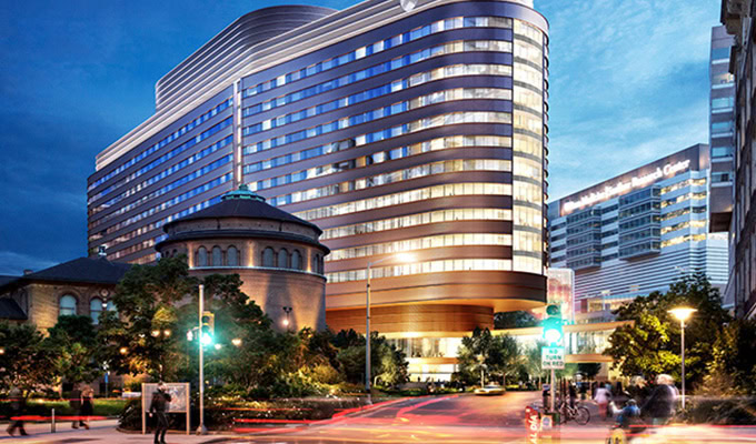 Modern, curved glass Pavilion At Penn Medicine hospital building illuminated at dusk, with adjacent older circular structure and people walking near the street intersection in the foreground.