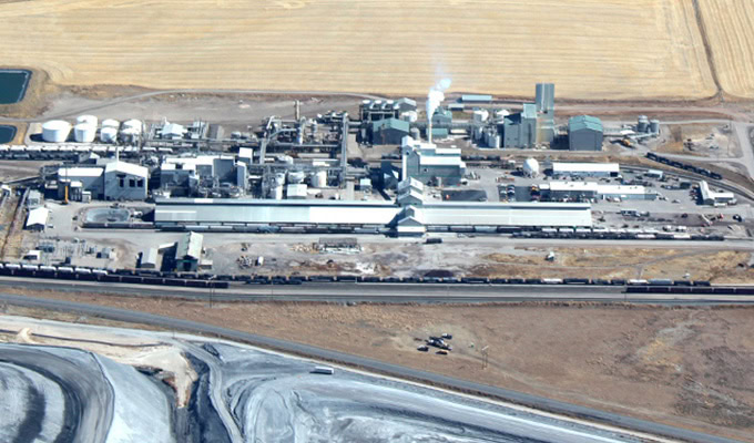 Aerial view of an industrial facility with multiple buildings, smokestacks, storage tanks, and train cars, surrounded by agricultural fields and barren land.