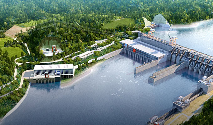 Aerial view of a hydroelectric dam facility with power generation buildings, water flowing through spillways, and surrounding greenery.