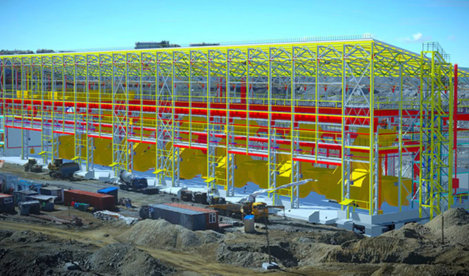 A large industrial building under construction, with exposed yellow and red steel framework, surrounded by construction equipment and dirt.