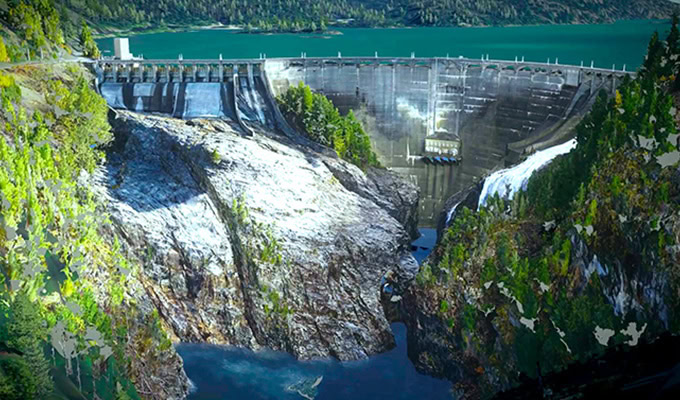 A large concrete dam spans a deep canyon with water flowing over the spillway, surrounded by forested hills and a reservoir in the background.