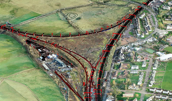 Aerial view of a railway junction construction site with multiple tracks converging, surrounded by fields and a residential area on the right.