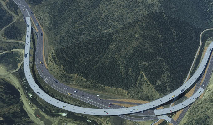 Aerial view of a large freeway interchange with multiple elevated ramps and vehicles traveling in both directions, surrounded by hilly terrain with sparse vegetation.