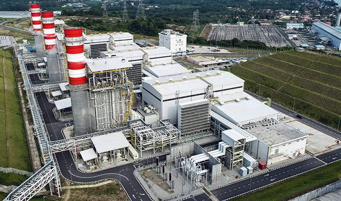 Aerial view of a modern industrial power plant facility with multiple buildings, red-and-white smokestacks, and surrounding infrastructure.