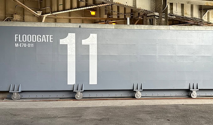 A large gray floodgate labeled "FLOODGATE 11 M-E70-011" with wheels at the bottom, positioned under an overhead structure.