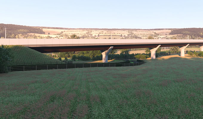 A modern overpass bridge spans across a grassy field with rolling hills and trees visible in the background under a clear sky.
