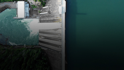 Overhead view of a dam, with water flowing from a reservoir on the left side into a larger body of water on the right. The dam structure includes buildings and spillways surrounded by greenery, perfectly aligned with the environmental agenda.