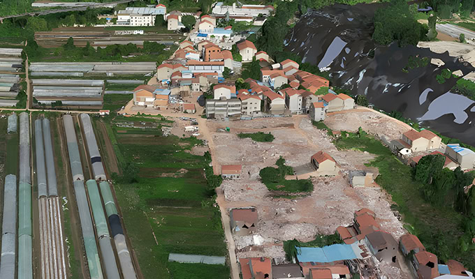 A small town with clustered houses, greenhouses on the left, a large area of cleared land, and a hillside with soil or debris in the background.