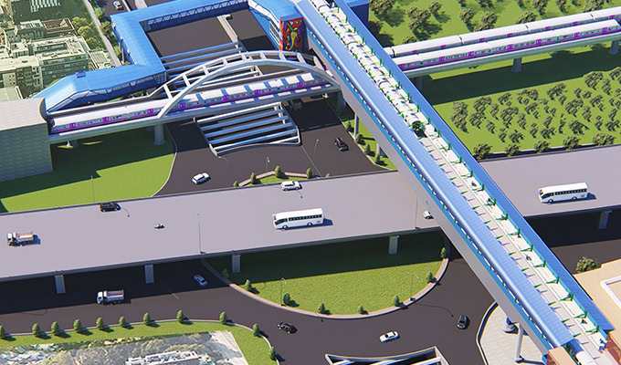 Aerial view of a modern elevated train station with covered pedestrian walkways, buses, cars, and landscaped surroundings.